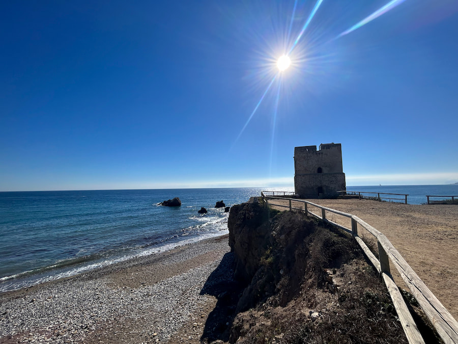Torre de vigilancia costera en Casares Costa, situada al borde de un acantilado con vista al mar Mediterráneo, bajo un cielo azul despejado y con el sol brillando intensamente; abajo se observa una playa de piedras.